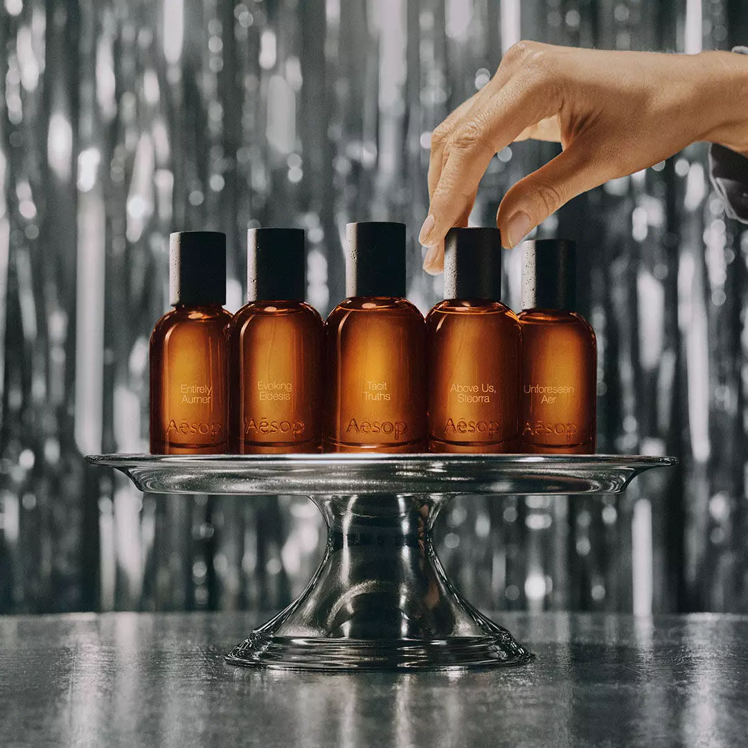 Two Aesop boxes, six small amber vials and a white nose-shaped object sit on a reflective ledge overlooking snow-covered mountains.