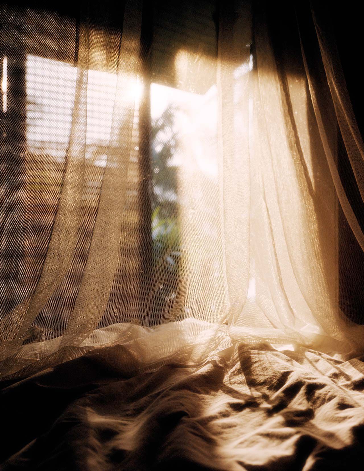 Sheer draped curtains over rumpled fabric near a sunlit window, showing warm beige tones, soft shadows, and translucent folds.