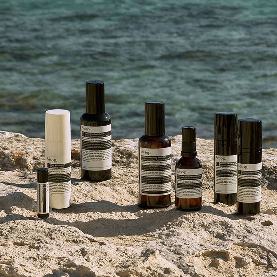 A curated selection of Aesop sun care protective partners displayed on a coastal rock by the sea.