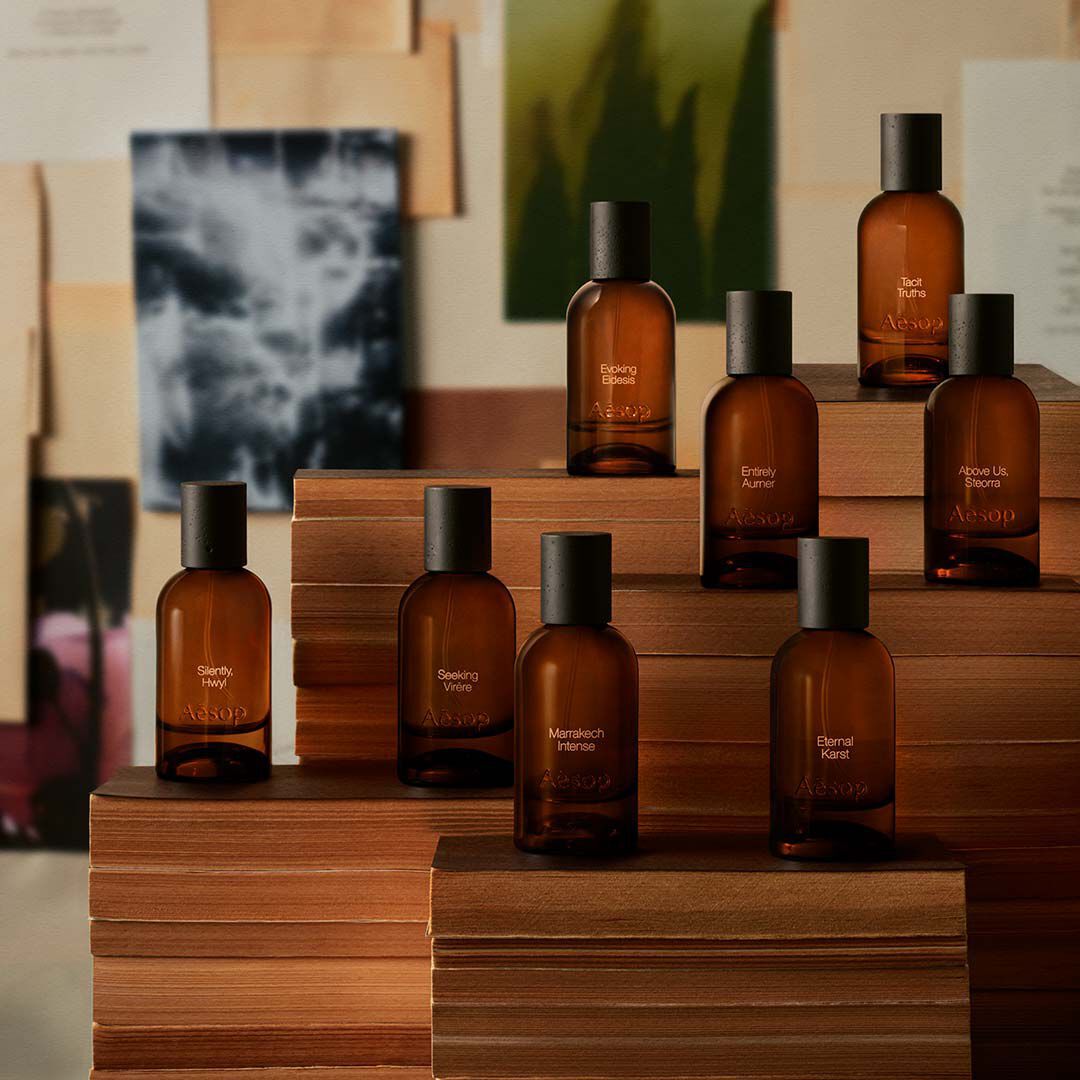 Collection of amber fragrance bottles arranged on wooden blocks before layered prints and papers in muted cream and charcoal tones.