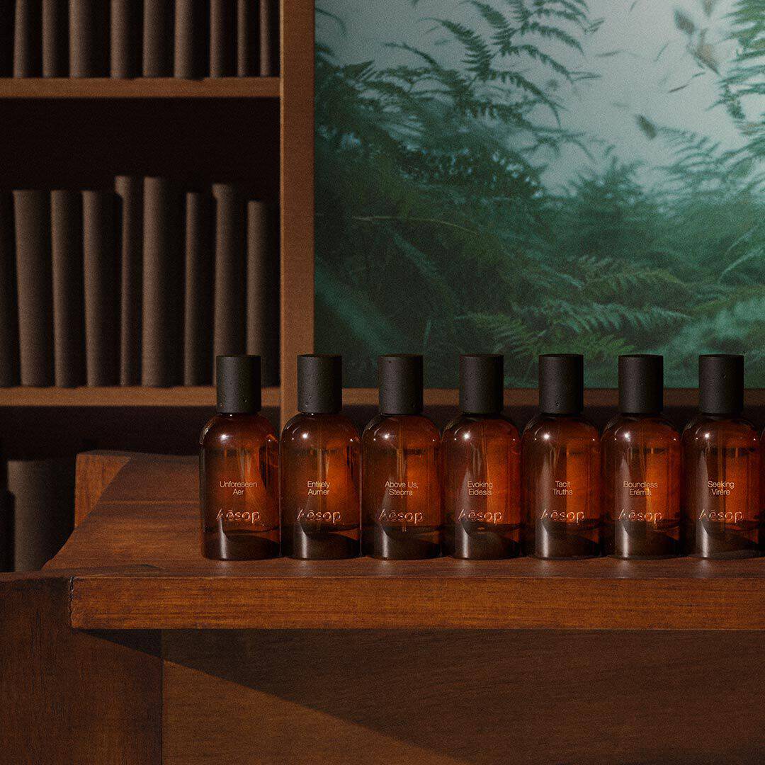 Row of amber Aesop fragrance bottles on a wooden table beside shelves and a green fern image.