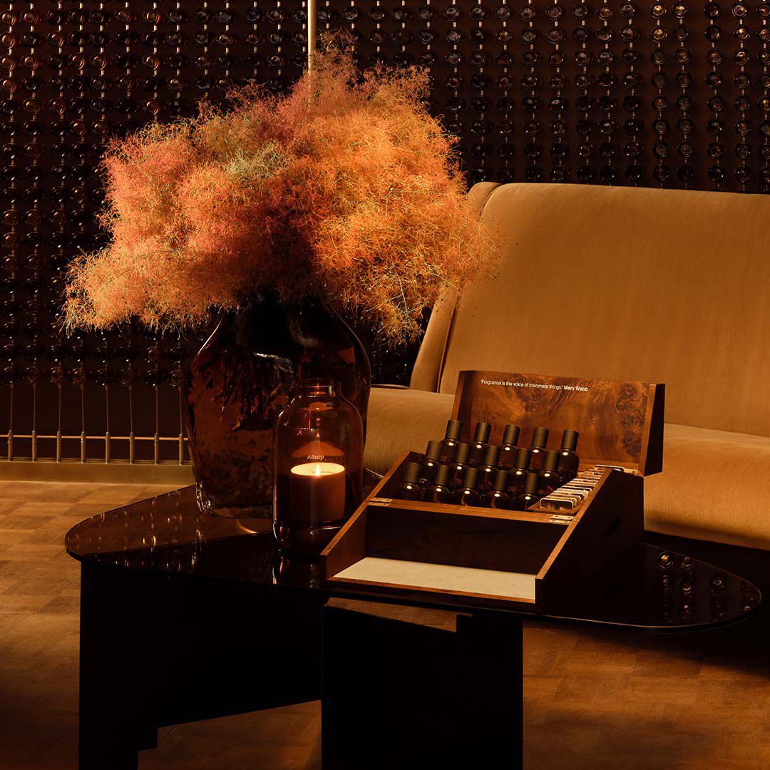 Amber-toned interior still life with a sofa, dark table, dried floral arrangement, and fragrance bottles displayed in an open presentation box.