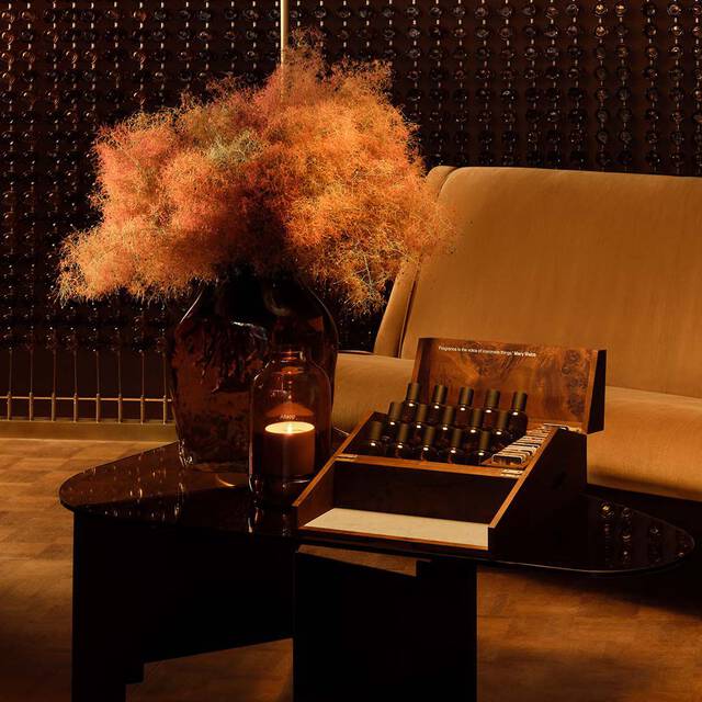 Amber-toned interior still life with a sofa, dark table, dried floral arrangement, and fragrance bottles displayed in an open presentation box.