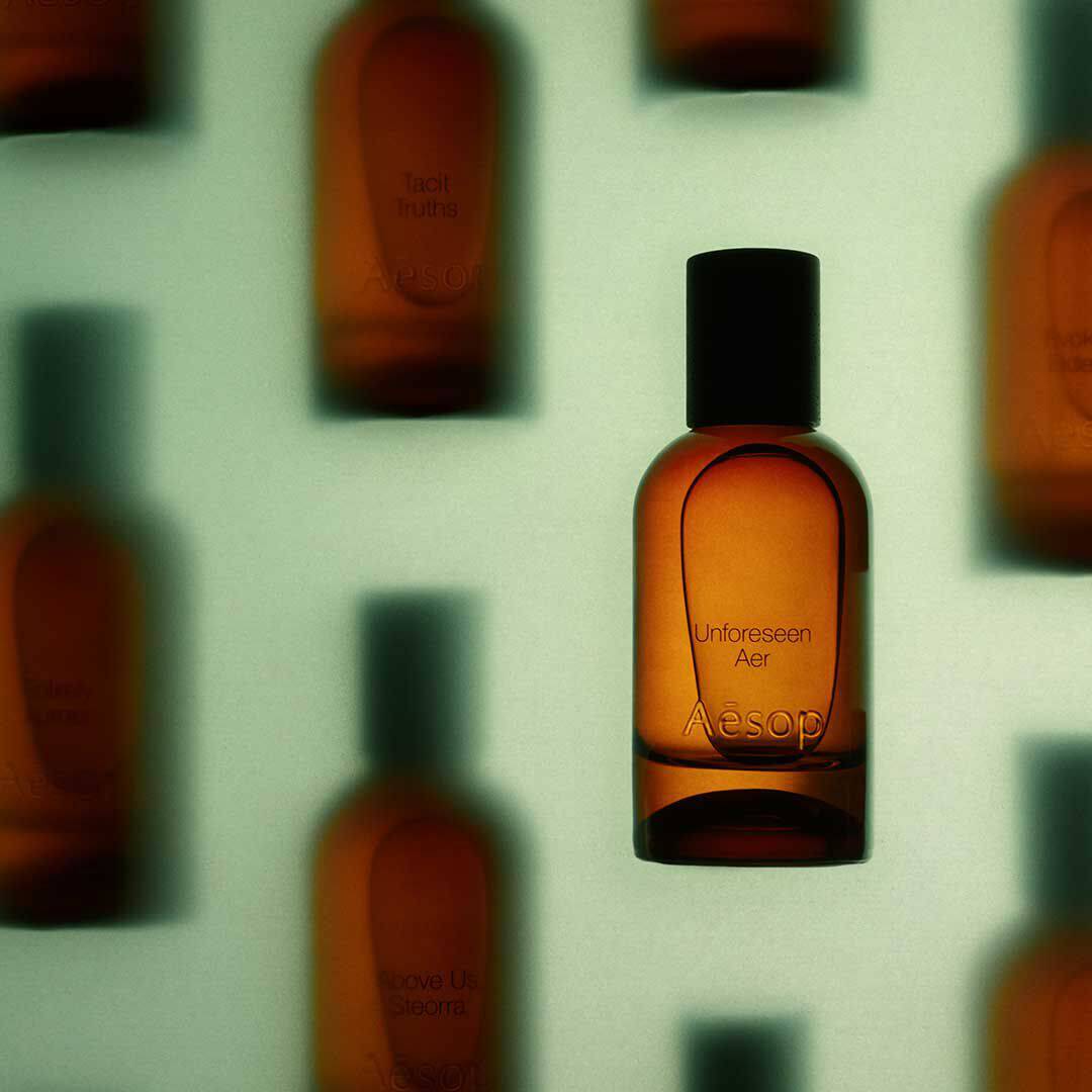 Aesop Unforeseen Aer amber fragrance bottle in focus before blurred amber bottles on a pale green background.