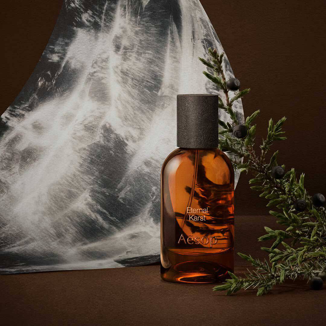 Amber Aesop fragrance bottle beside rosemary sprigs and a monochrome water image on a dark brown surface.