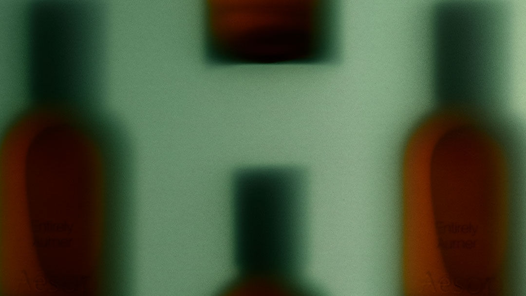 Soft-focus amber fragrance bottles with dark caps against a pale green backdrop, creating a blurred repeating horizontal pattern.