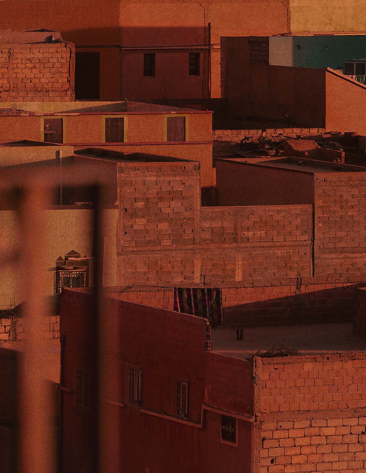 Terracotta city rooftops and walls in warm rust-red light, with blocky architecture, shadowed facades, and textured masonry surfaces.