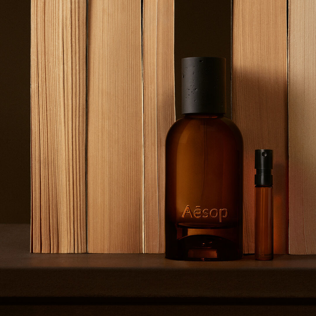 Amber Aesop bottle and slim vial placed between vertical pale wood boards, with deep brown shadows and a clean linear composition.