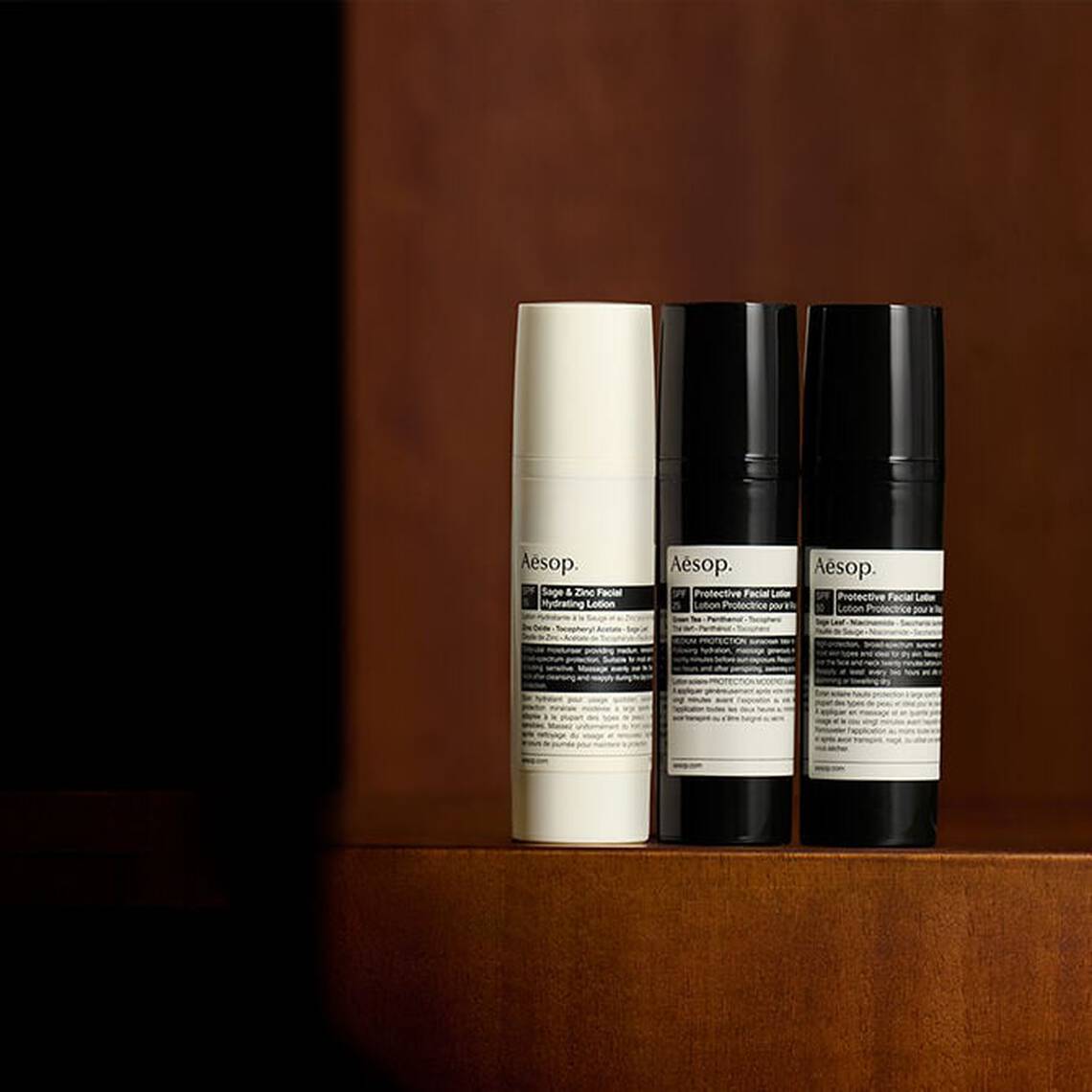 A curated selection of Aesop sun protection formulations standing on a dark wooden surface in a softly lit setting.
