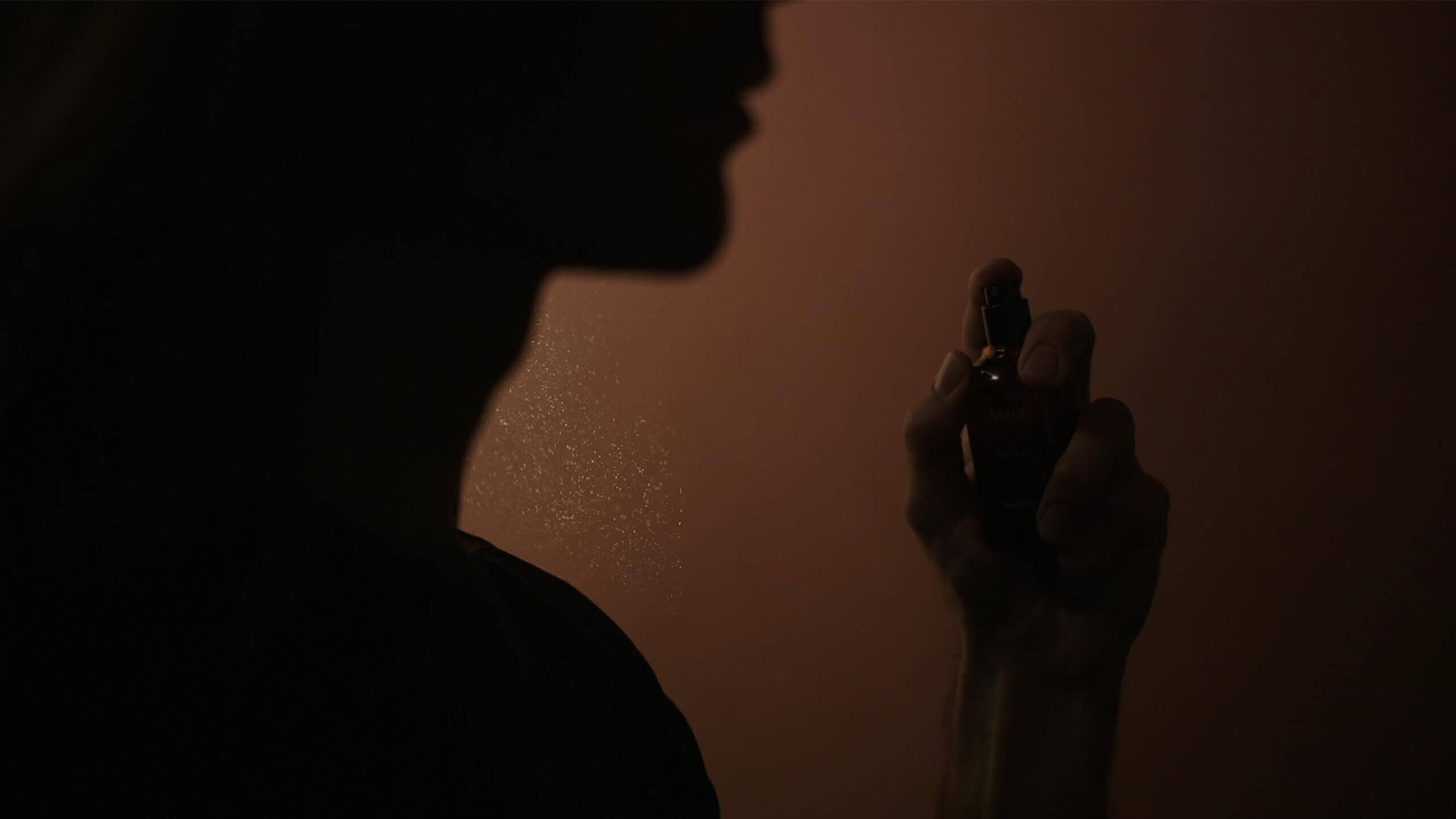 Silhouette of a person holding a small fragrance bottle near their face against a warm amber background.