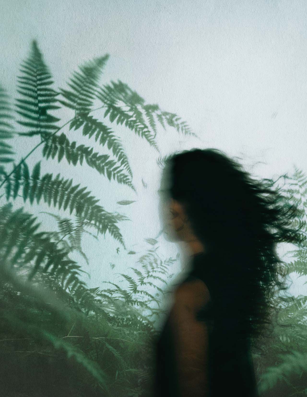 Blurred profile of a person with dark hair among green ferns, set against misted glass and cool diffused natural light.