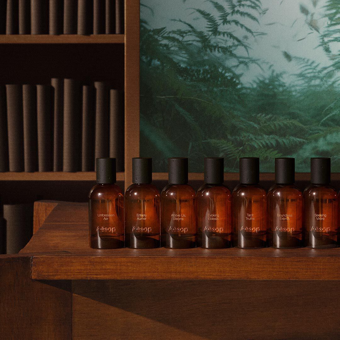 Row of amber glass fragrance bottles with black caps on stepped wooden shelving, with books and fern imagery in the background.